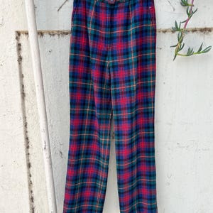 May include: A pair of vintage plaid pants in a classic tartan pattern. The pants feature a vibrant mix of red, green, blue, and yellow lines, creating a bold and eye-catching design. The pants are hanging on a clear hanger.
