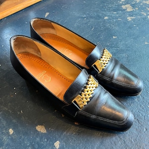 Puede incluir: Un par de mocasines de cuero negro con detalles de cadena dorada en la pala. Los zapatos tienen una puntera redondeada y un tacón bajo. El interior presenta una plantilla de color beige. Los zapatos se exhiben sobre una superficie azul oscuro.