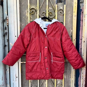 May include: Red hooded jacket with white trim and a white interior. The jacket features a zig-zag stitch design and two front pockets. The jacket is buttoned up and hanging on a black hanger.