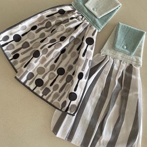 May include: Two kitchen towels with hanging loops. One towel has a grey and white striped pattern. The other towel has a white background with a pattern of black and grey kitchen utensils. Both towels have a small pocket with a button closure.