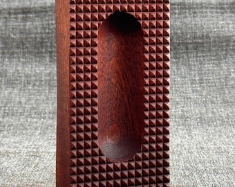 May include: A dark red wooden rectangular box with a carved oval-shaped indentation in the center. The box has a textured surface with a pattern of small triangles.