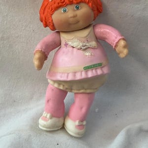 May include: A small plastic doll with bright orange hair styled with two white bows. The doll wears a pink dress with a white collar and a white bow detail on the chest, along with pink leggings and white shoes. A green label is visible on the dress.