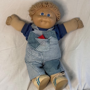 May include: A vintage doll with a tan face, blue eyes, and light brown yarn hair. The doll wears a blue shirt, denim overalls, and beige shoes with blue stripes. A red fabric is visible in the overalls pocket.