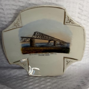 May include: A decorative, off-white ceramic dish with a painted image of the Illinois Central Bridge in Cairo, Illinois. The bridge is depicted over water with a sunset sky. The text "THE ILLINOIS CENTRAL BRIDGE, CAIRO ILL. 5 MILES LONG." is printed below the image.
