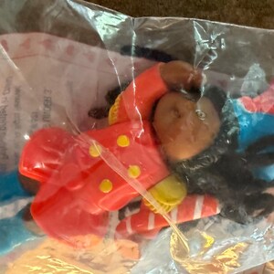 May include: A colorful toy clown figure in a clear plastic bag. The clown has a brown face, black hair, a red jacket with yellow buttons, and blue shoes. The clown's hat is blue and red.