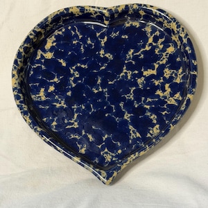 May include: A heart-shaped ceramic dish featuring a dark blue and cream-colored sponge-painted or speckled pattern. The dish has a raised rim and a smooth, glossy finish, with the blue and cream colors blended in an irregular, mottled design.