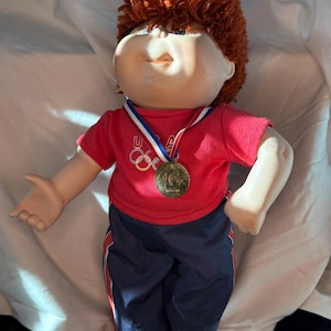 May include: A Cabbage Patch Kid doll wearing a red t-shirt with the Olympic rings logo and navy blue track pants with red and white stripes. The doll has red hair and a gold medal.