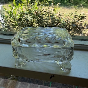 May include: A clear, vintage glass trinket box with a lid. The box has a textured, swirling design on the lid and sides, and rests on four small feet. The box is likely used for storing jewelry or small items.