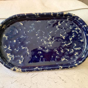 May include: An oval-shaped ceramic tray featuring a deep navy blue glaze with a speckled, cream-colored pattern across the surface. The tray has a raised rim and a glossy, reflective finish, showcasing a textured, uneven surface throughout the interior.
