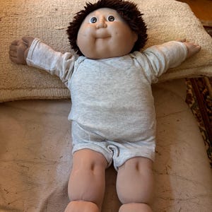 May include: A doll with a round face, dimpled cheeks, and short brown yarn hair. The doll has a light skin tone, dark eyes, and is wearing a light gray long-sleeved onesie with a simple round neckline and short legs that cover the torso.