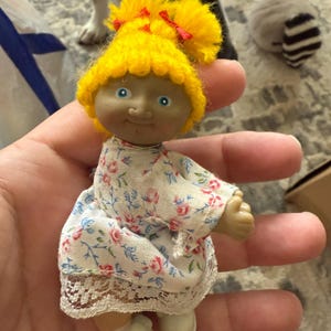 May include: A small plastic doll with yellow yarn hair, a white dress with pink flowers, and white shoes. The doll is smiling and has blue eyes.