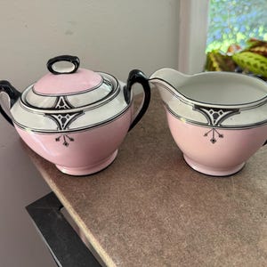May include: A pink and white ceramic sugar bowl with a lid and a creamer. Both pieces have black handles and decorative black and white geometric patterns. The sugar bowl has a black oval handle on the lid. The set is on a brown surface.