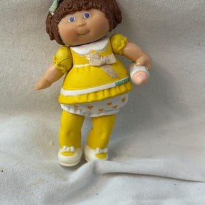 May include: A Cabbage Patch Kids doll with brown hair in pigtails, blue eyes, and a light green bow. The doll wears a yellow dress with a white collar, a beige ribbon, and a white skirt with yellow hearts, yellow leggings, and white shoes.