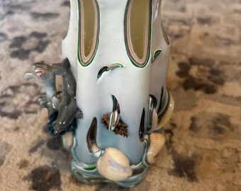 May include: A white and blue carved candle with a seashell and dolphin design. The candle is made of layered wax and has a hollow center.