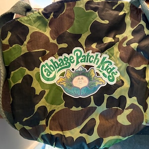 May include: A camouflage-patterned shoulder bag featuring the Cabbage Patch Kids logo. The bag has a green strap and a repeating pattern of brown, black, and green shapes. The logo is a green and yellow emblem with the words "Cabbage Patch Kids".