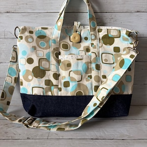 May include: A patterned tote bag with a navy blue base and a cream-colored top featuring blue and brown geometric shapes. The bag has a matching adjustable shoulder strap and a wooden button closure. The bag is hanging on a white hook.