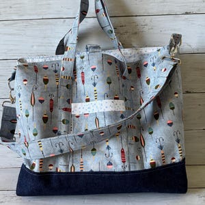May include: A large tote bag with a light blue background and a pattern of colorful fishing bobbers. The bag has a dark blue denim base and a white accent stripe. It features both shoulder straps and a crossbody strap.