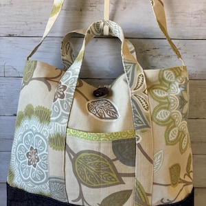 May include: A beige tote bag with a floral pattern in green, brown, and gray. The bag has a dark blue denim base, a front pocket, and two sets of handles: short handles and a long shoulder strap.