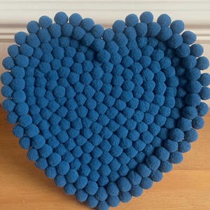 May include: A heart-shaped decorative item made of small, round, blue pom-poms. The pom-poms are densely packed together, creating a textured surface. The item is likely a decorative piece for a home or event.