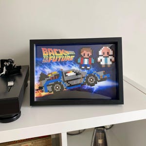 Puede incluir: Una pieza de arte de píxeles enmarcada que presenta la máquina del tiempo DeLorean de la película Regreso al futuro. El arte también incluye representaciones de píxeles de Marty McFly y Doc Brown.