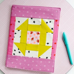 Peut inclure: Une couverture de carnet matelassée avec un motif patchwork. Le centre présente un motif géométrique jaune et blanc avec un carré floral rose. La bordure est rose avec des motifs d'étoiles et de croix blanches. Un stylo turquoise repose sur le tapis de coupe rose.