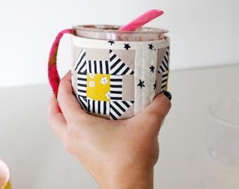 Può includere: Un rivestimento per tazza in tessuto fatto a mano con un design patchwork. Il rivestimento presenta uno sfondo beige con motivi geometrici bianchi e neri, quadrati gialli e piccoli accenti di stelle nere. Un cucchiaio rosa è all'interno della tazza.