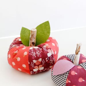 May include: Two handmade fabric pumpkins with patchwork designs. The larger pumpkin is red and pink with a green felt leaf on top. The smaller pumpkin is burgundy and black with a pink felt leaf on top. Both pumpkins have a wooden stem.
