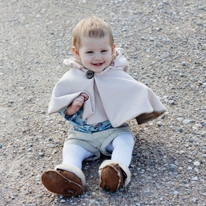 May include: A young child wearing a light pink cape with a brown button sits on a gravel surface. The child is wearing brown shoes and a blue and white patterned shirt.