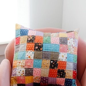 Może przedstawiać: Poduszka patchworkowa z różnorodnych kolorowych tkanin w kwadratowym wzorze. Poduszka leży na różowym fotelu.