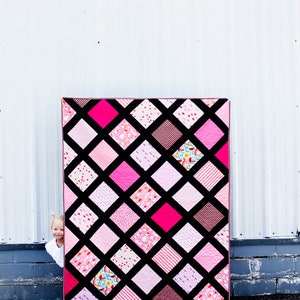 May include: A pink and black quilt with a diamond pattern. The quilt is made of various pink fabrics with different patterns. The quilt is leaning against a white wall.