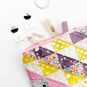 May include: A quilted zippered pouch with a pink, purple, yellow, and white patchwork design. The pouch is open and lying on a white surface with various accessories, including earrings, a hair clip, and a bottle of perfume.