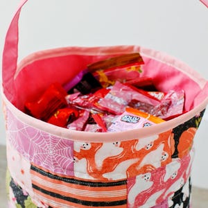 Puede incluir: Cubo de tela rosa para pedir dulces con un dise&ntilde;o de patchwork con fantasmas, telara&ntilde;as y las palabras "boo". El cubo est&aacute; lleno de caramelos.