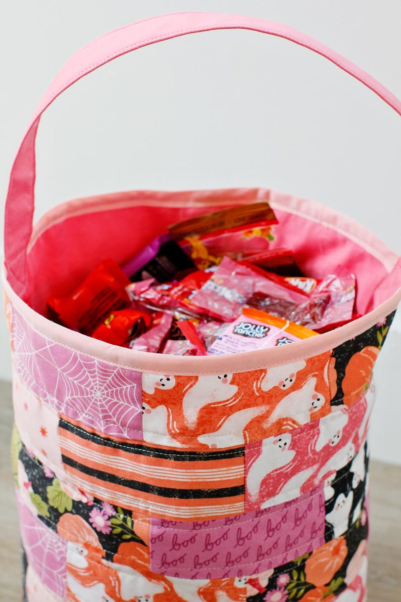 Puede incluir: Cubo de tela rosa para pedir dulces con un dise&ntilde;o de patchwork con fantasmas, telara&ntilde;as y las palabras "boo". El cubo est&aacute; lleno de caramelos.