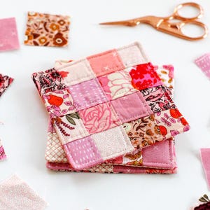 Puede incluir: Una pila de cuatro posavasos de tela de patchwork rosa y rojo con motivos florales. Los posavasos están hechos de cuadrados de diferentes telas cosidos juntos.