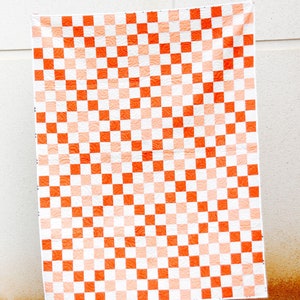 May include: A large quilt with a checkered pattern in shades of orange and white. The quilt is hanging on a white wall and the bottom edge is resting on green grass.