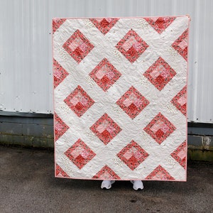 May include: A white quilt with a pink and white floral pattern. The quilt is made up of squares with a diamond pattern in the center. The quilt is hanging on a white wall.