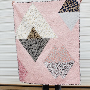 Pode incluir: Uma colcha rosa com um padrão geométrico de diferentes tecidos com estampas de animais. A colcha é composta por quatro triângulos grandes, cada um com um padrão diferente. A colcha está sendo segurada pelas mãos de uma pessoa.