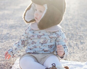 May include: A baby wearing a brown faux fur hat with a pink bow and a floral print blouse, sitting on a white blanket.