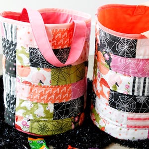 Puede incluir: Dos bolsas de tela de Halloween para pedir dulces de color rosa y naranja con dise&ntilde;os de patchwork que incluyen fantasmas, ara&ntilde;as y telara&ntilde;as. Las bolsas tienen asas rosas y est&aacute;n llenas de caramelos.