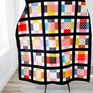 Könnte beinhalten: Ein farbenfroher Patchwork-Quilt mit schwarzem Rand, präsentiert auf einem Holzständer. Der Quilt zeigt Quadrate aus verschiedenen gemusterten Stoffen in Rot-, Rosa-, Gelb-, Blau- und Orangetönen. Der Quilt ist ca. 180 cm x 200 cm groß.