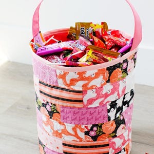 Puede incluir: Un cubo de tela rosa para pedir dulces con un dise&ntilde;o de patchwork que presenta fantasmas, calabazas y las palabras "boo" en rosa y negro. El cubo est&aacute; lleno de varios dulces, incluyendo piruletas, barras de chocolate y caramelos gomosos.
