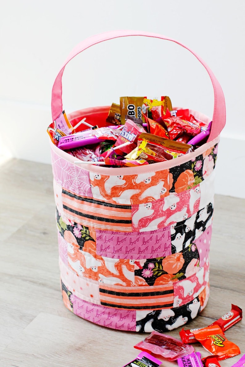 Puede incluir: Un cubo de tela rosa para pedir dulces con un dise&ntilde;o de patchwork que presenta fantasmas, calabazas y las palabras "boo" en rosa y negro. El cubo est&aacute; lleno de varios dulces, incluyendo piruletas, barras de chocolate y caramelos gomosos.