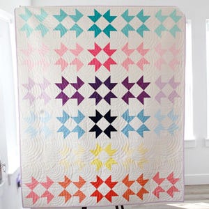 Puede incluir: Una colcha colorida con un patrón de estrella en forma de estallido en tonos de rosa, morado, azul, amarillo y naranja. La colcha se exhibe en un soporte en una habitación con paredes blancas y pisos de madera.