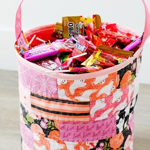 Może przedstawiać: Różowy materiałowy kubełek na cukierki z patchworkowym wzorem przedstawiającym duchy, pająki i napis "boo" w kolorze różowym. Kubełek jest wypełniony cukierkami i lizakami.