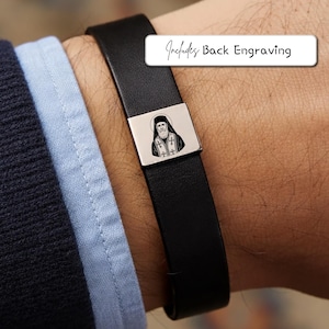 Può includere: Bracciale in pelle nera con chiusura rettangolare argentata con incisione di una figura religiosa. Il testo "Includes Back Engraving" è visualizzato su un'etichetta rettangolare bianca.