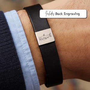 Può includere: Bracciale in pelle nera con chiusura rettangolare color argento con design di pesce gatto inciso. Il testo "Includes Back Engraving" è visualizzato su un'etichetta bianca. Il bracciale è indossato al polso.