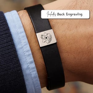 Może przedstawiać: Czarna skórzana bransoletka ze srebrnym zapięciem. Zapięcie ma motyw naukowy i tekst "Includes Back Engraving". Bransoletka jest noszona na nadgarstku.