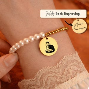 Può includere: Un braccialetto color oro con un design di perle e perline. Il braccialetto presenta un ciondolo rotondo con un'illustrazione in bianco e nero di una persona che tiene in braccio un bambino. Il ciondolo ha anche il testo "je t'aime mon amour". Il testo "Includes Back Engraving" è anche visibile.