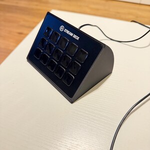 Könnte beinhalten: Schwarzes Stream Deck mit 15 programmierbaren Tasten. Das Gerät hat das Stream Deck-Logo und den Text oben. Ein schwarzes Kabel erstreckt sich von dem Gerät. Das Gerät befindet sich auf einer weißen Oberfläche.