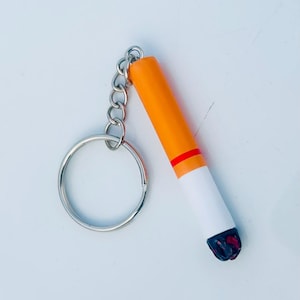 May include: A novelty keychain shaped like a cigarette. The keychain features a silver metal split ring and chain. The cigarette is orange, white, and black with a red band.